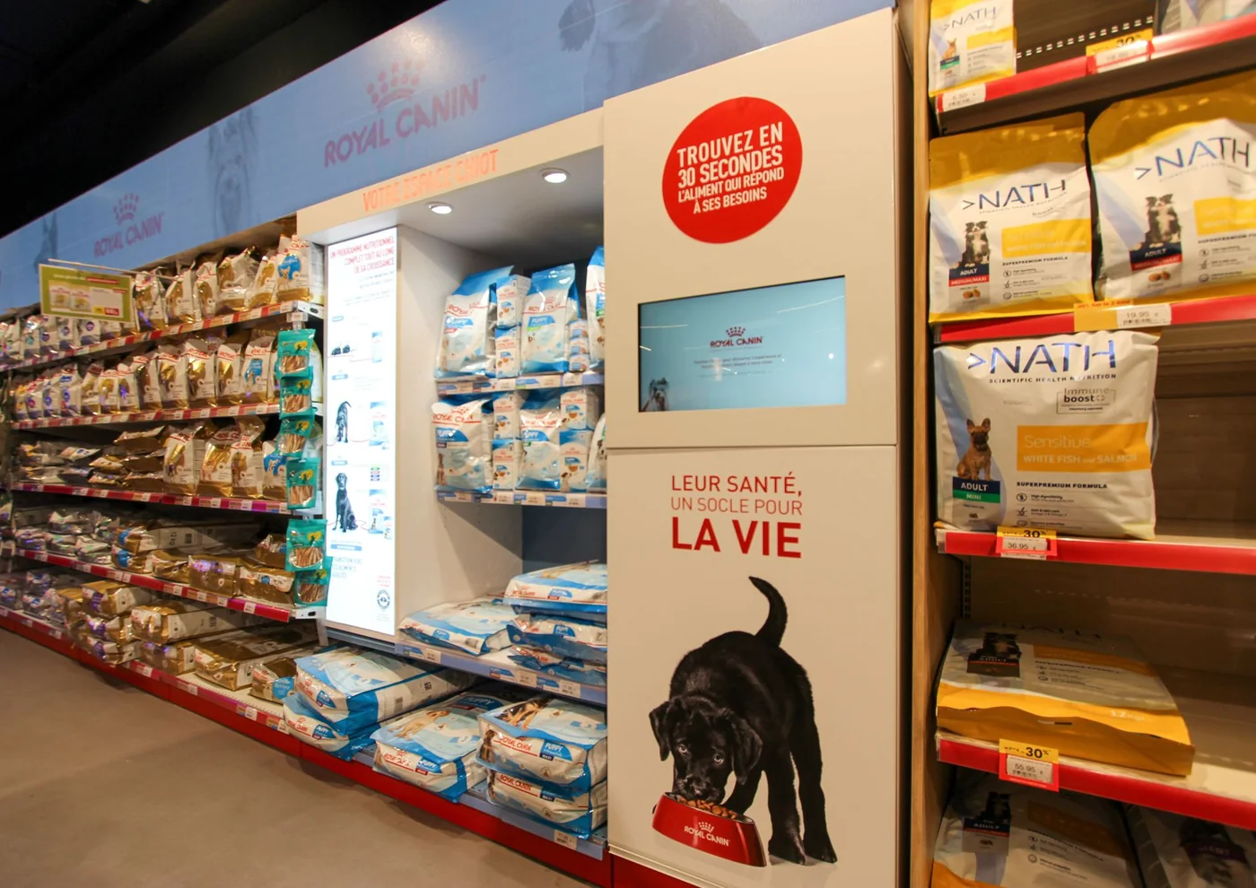 Borne tactile Royal Canin intégrée au linéaire d'une animalerie : « Trouvez en 30 secondes l'aliment qui répond à ses besoins », entre les rayons de sacs de croquettes.